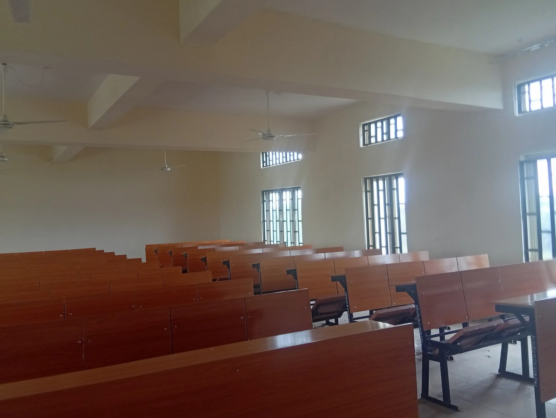 An empty classroom with rows of wooden desks, side windows and ceiling fans
